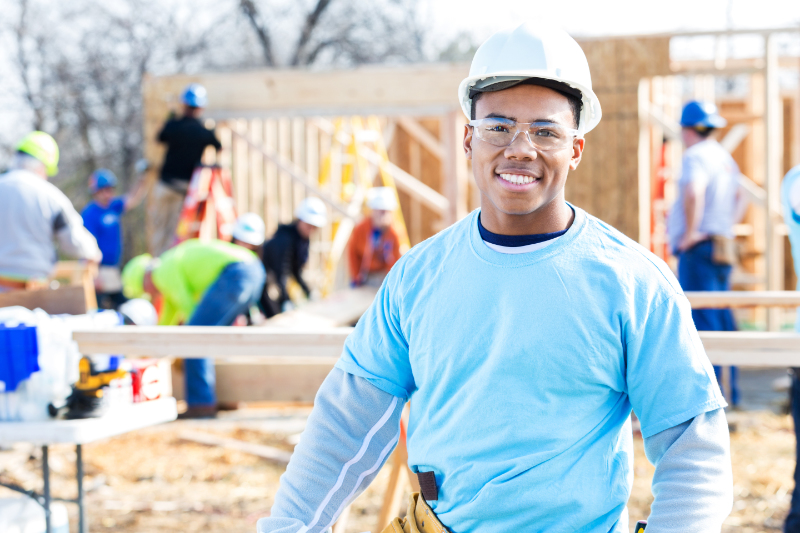 Schedule Your Annual Furnace Inspection Now - Handsome African American construction foreman or building contractor stands in front of home that is being built for charity. Volunteers are working on the home in the background. He hsi wearing a hard hat, safety glasses and volunteer t-shirt.