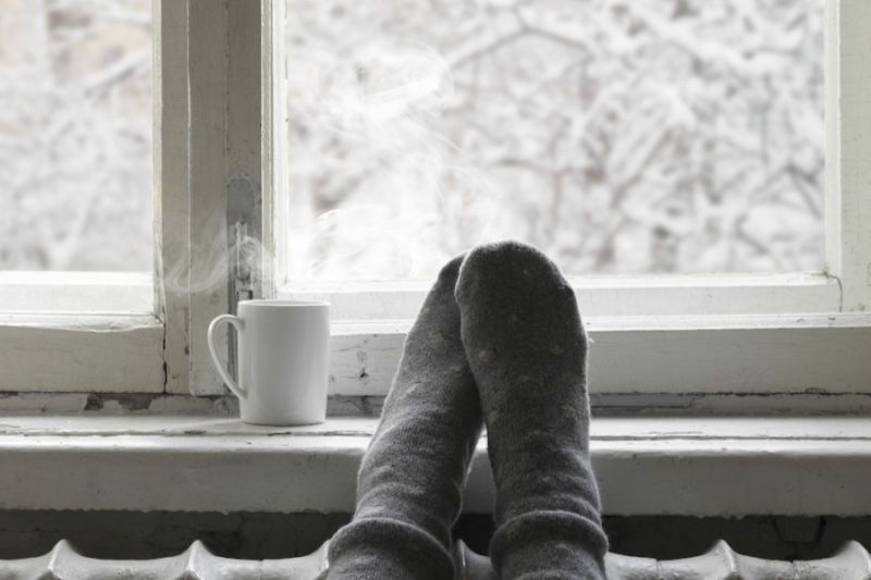 How Do I Keep My Heat During Extreme Cold Weather? Image shows a person's feet resting on a window sill with a cup of hot drink.