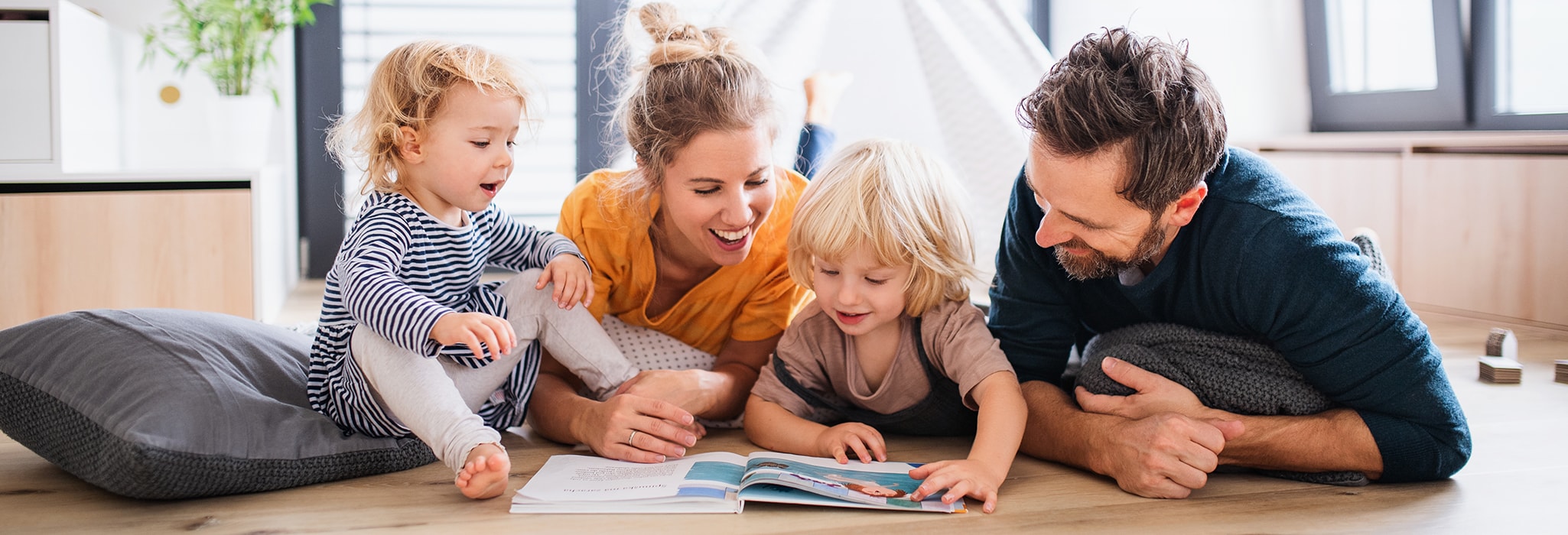 Front view of young family with two small children indoors in bedroom reading a book.